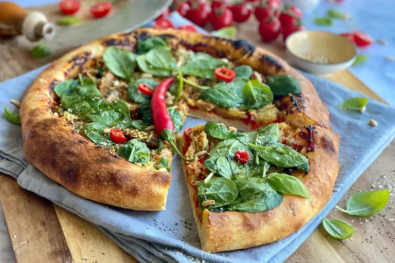 Nahaufnahme einer Pizza mit frischem Spinat, roten Chilischoten und Pinienkernen auf einem Holzbrett, umgeben von Tomaten.