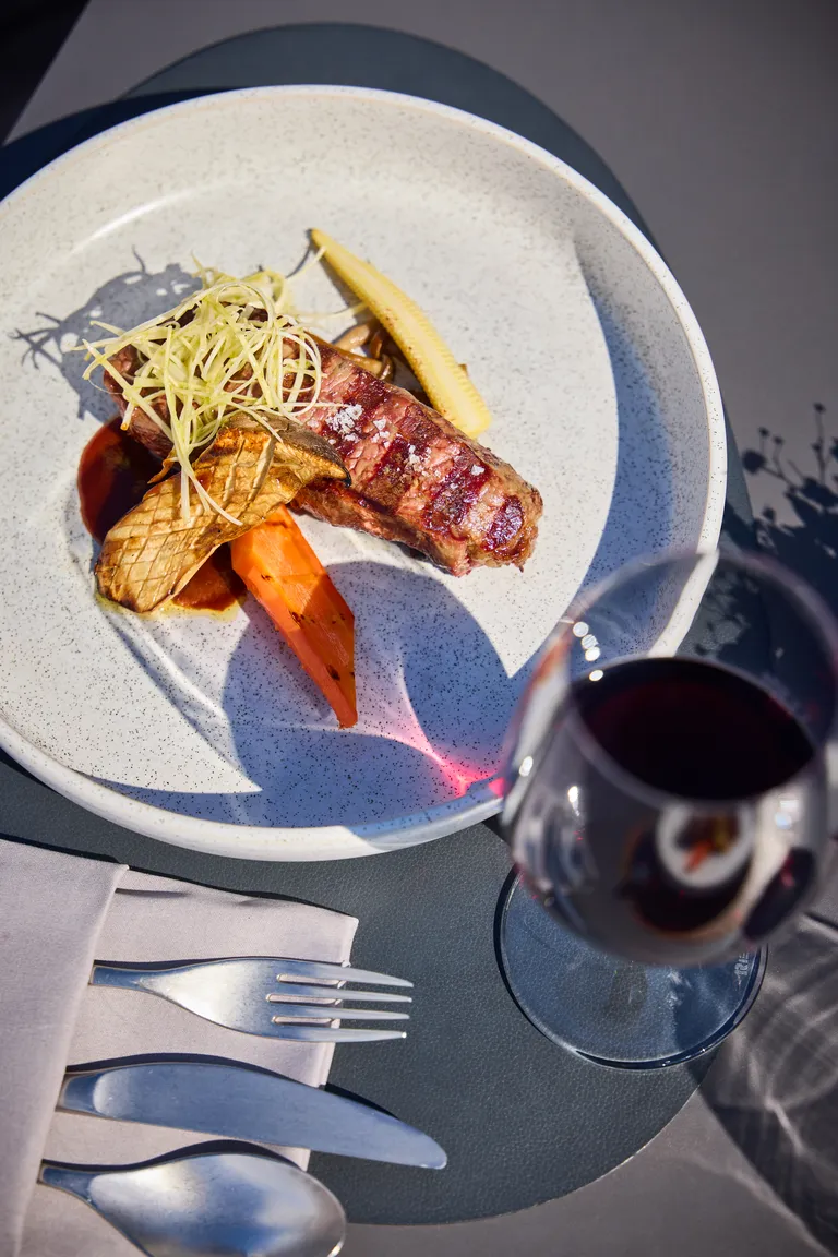 Gegrilltes Beiried vom Angus mit Kukuruz, Pilzen und Gemüse auf einem Teller, dazu ein Glas Rotwein