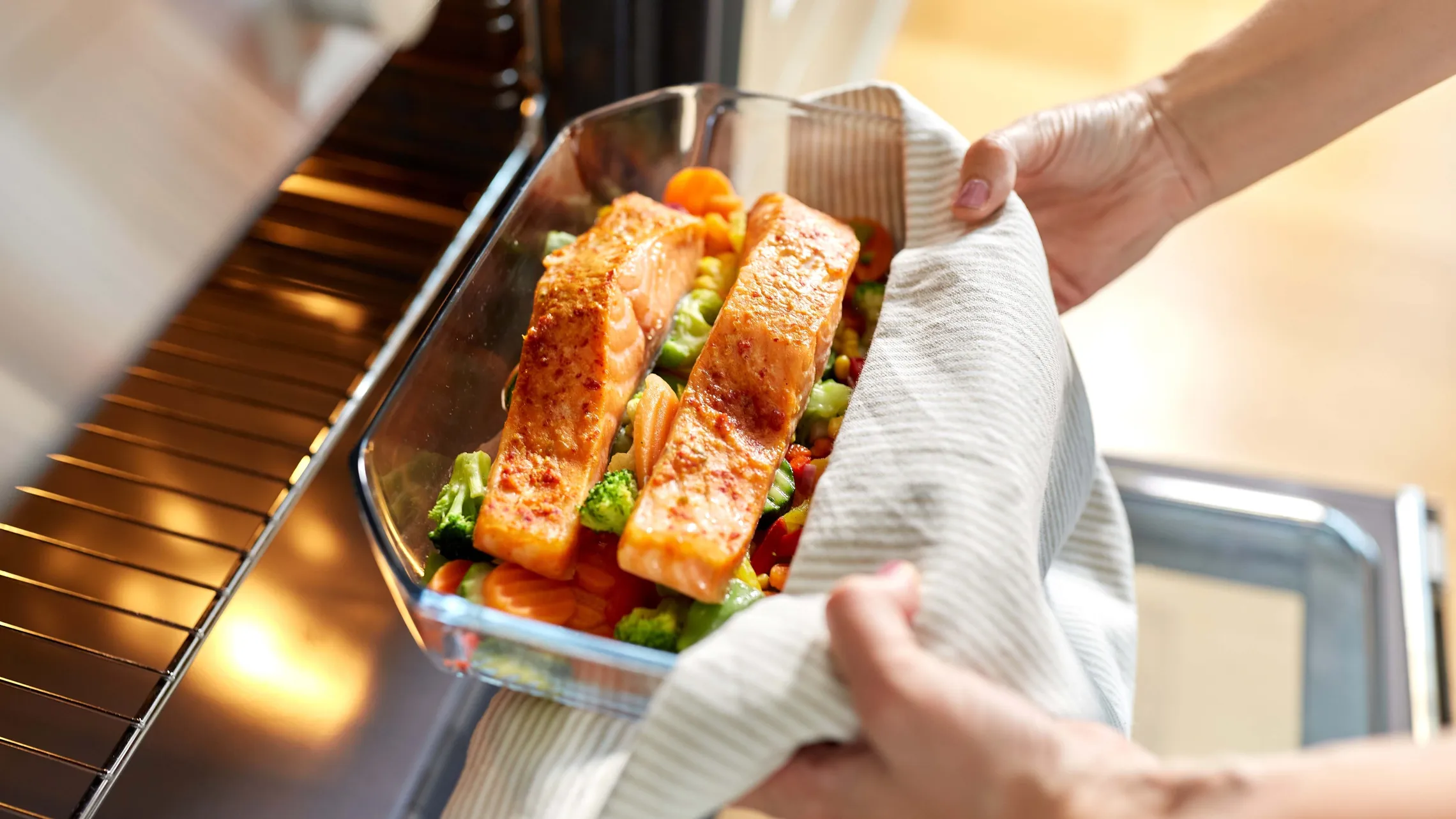 Zwei Lachsfilets auf geröstetem Gemüse in einer Glasform, die mit einem Tuch aus dem Ofen genommen werden.