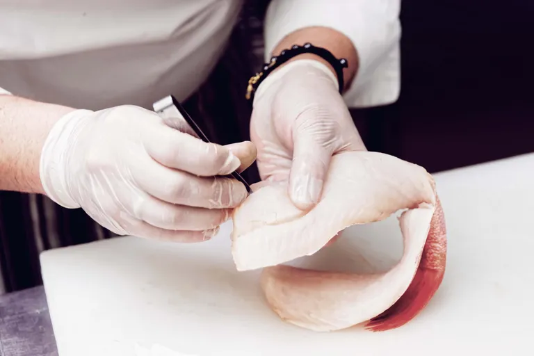 Ein Koch mit Handschuhen, der auf einem Schneidebrett mit einer Pinzette ein rohes Fischfilet ausbeint.