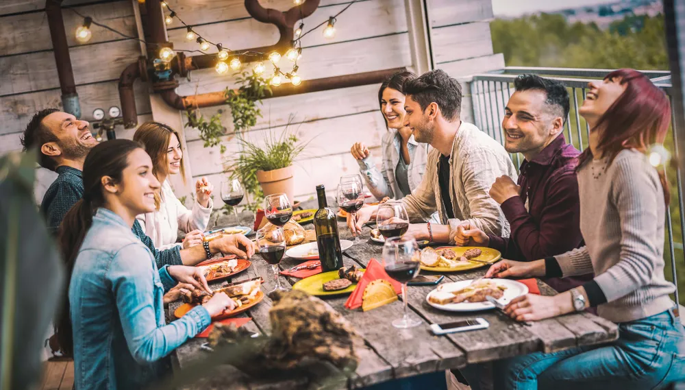 Eine Gruppe von Freunden, die an einem rustikalen Holztisch im Freien bei Lichterketten über ihren Köpfen essen und trinken, lachen und sich unterhalten.