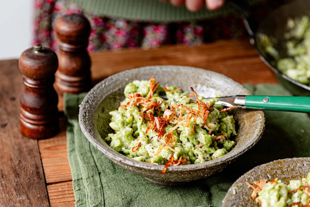 Eine Schüssel cremige grüne Spinatspätzle mit knusprig gebratenen Zwiebeln, serviert auf einer grünen Serviette neben Pfeffermühlen auf einem Holztisch.