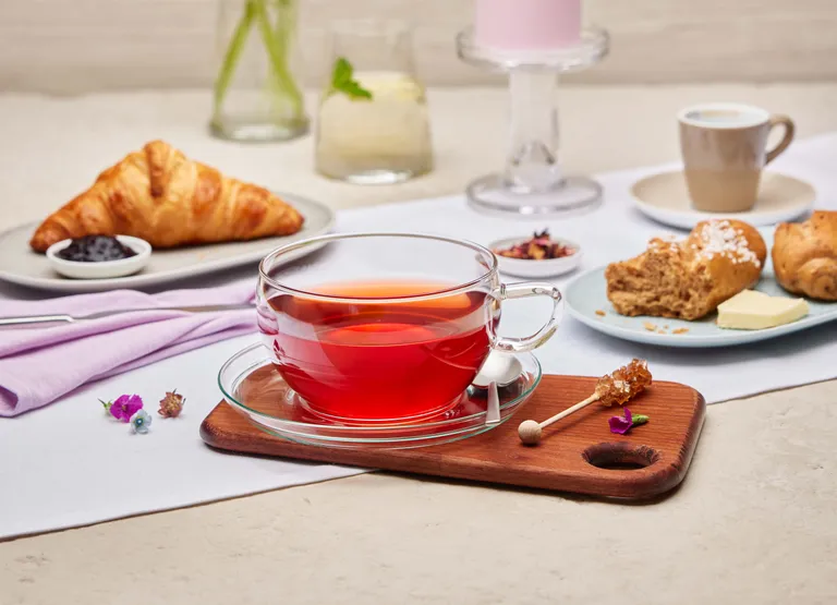 Ein Frühstückstisch mit einem Glas rotem Tee, Croissants, Scones, einer rosa Kerze und Blumen in einer Vase auf einer hellen, strukturierten Oberfläche.