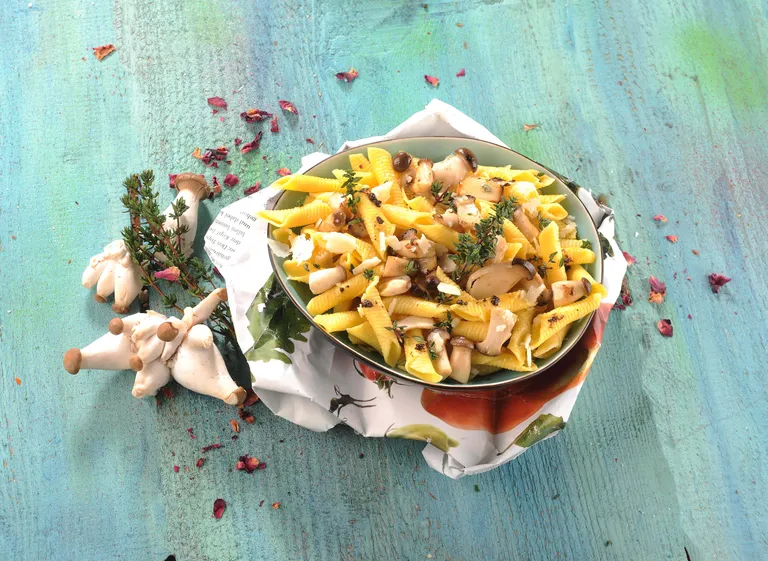 Eine Schüssel Pasta mit Pilzen, Kräutern und geriebenem Käse auf einem blauen Holztisch, umgeben von verstreutem Thymian, Pilzen und getrockneten Blütenblättern.