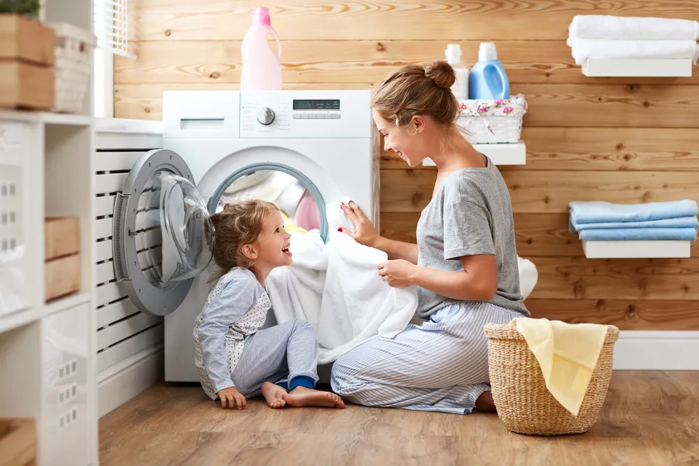 Mutter und Tochter genießen gemeinsam die Zeit beim Wäschewaschen, während das Kind in einem hellen und aufgeräumten Waschraum in die Waschmaschine hineinschaut.