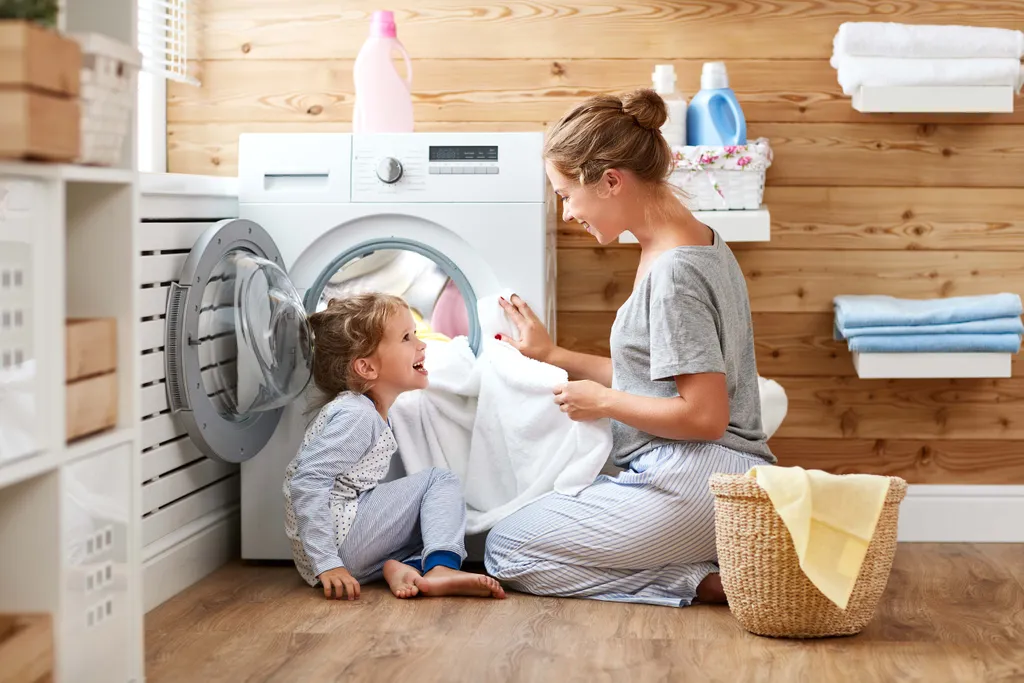 Mutter und Tochter genießen gemeinsam die Zeit beim Wäschewaschen, während das Kind in einem hellen und aufgeräumten Waschraum in die Waschmaschine hineinschaut.