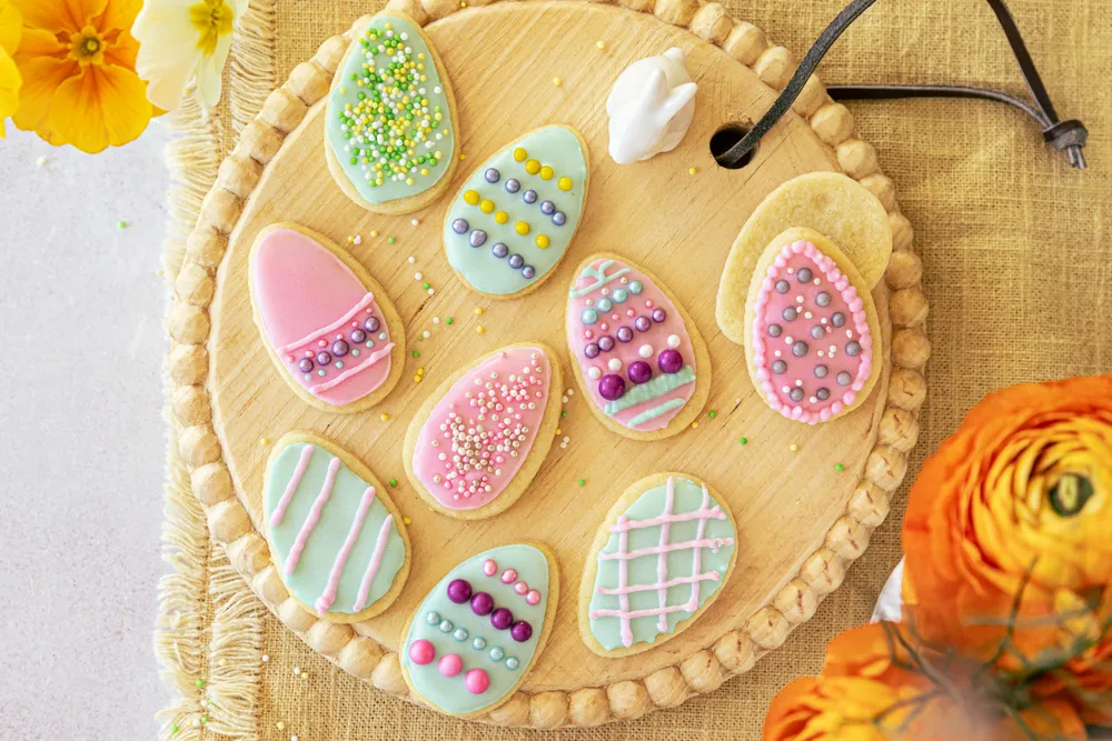 Bunte, osterei-förmige Kekse mit Zuckerguss und Streuseln auf einem Holzbrett, umgeben von orangefarbenen und gelben Blumen.