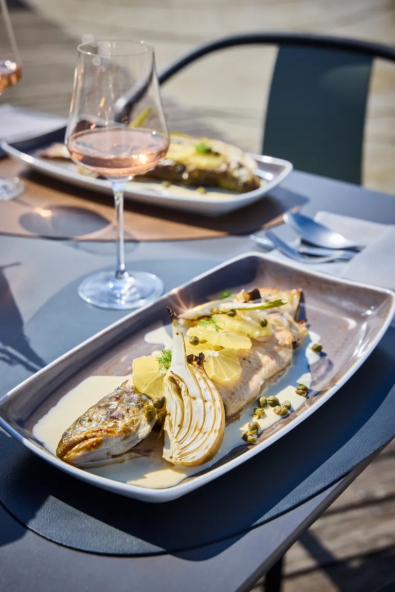 Gegrillter Saibling mit Fenchel, Zitronenscheiben und Kapern auf einer länglichen Platte, dazu ein Glas Wein