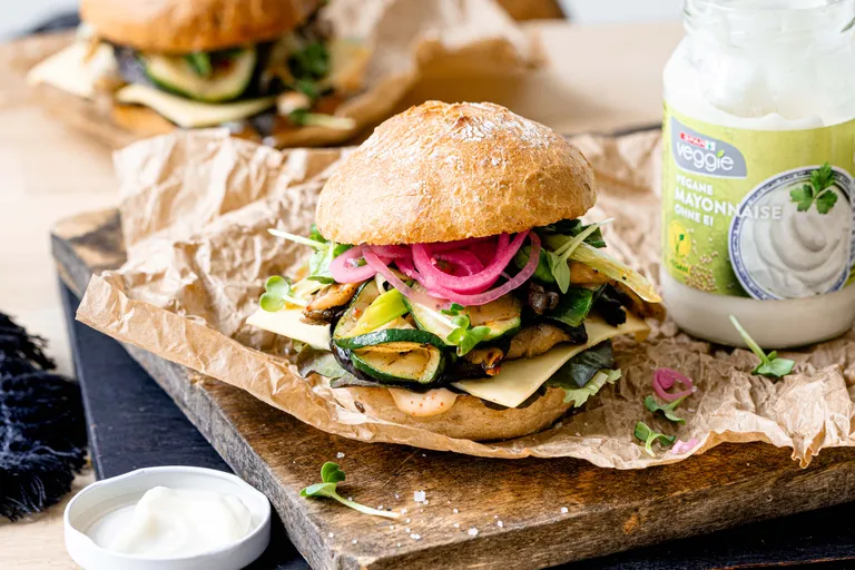 Veganer Burger mit gegrillter Zucchini, Käse, eingelegten Zwiebeln und Blattgemüse auf einem Brötchen, daneben ein Glas vegane Mayonnaise auf einem Holzbrett.
