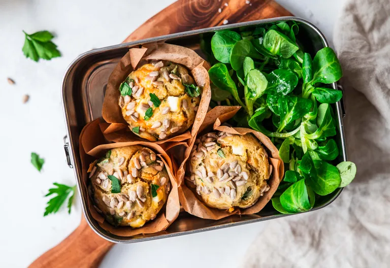 Drei herzhafte Muffins mit Samen und Kräutern in einer Brotdose, daneben frischer grüner Salat.