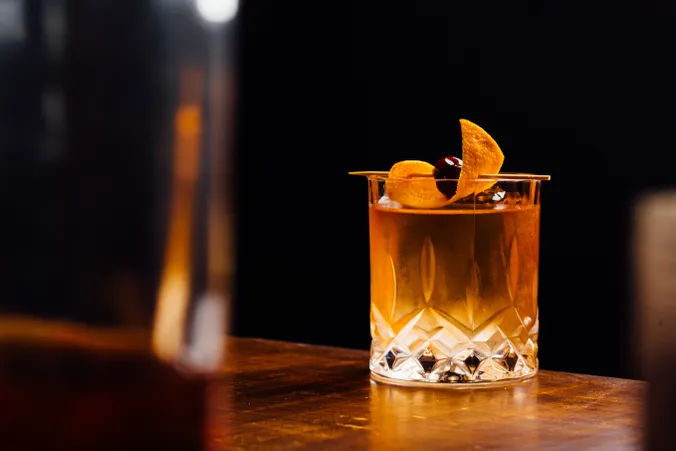 Ein Kristallglas, gefüllt mit einem orangefarbenen Cocktail, garniert mit einer Orangenschale und einer Kirsche, auf einer Holzunterlage vor einem dunklen Hintergrund.