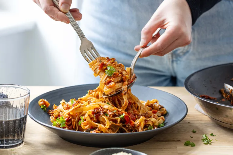 Eine Person, die auf einem blauen Teller Pasta mit Tomatensoße und Kräutern serviert, wobei sie Gabel und Löffel benutzt, daneben steht ein Glas Wasser.