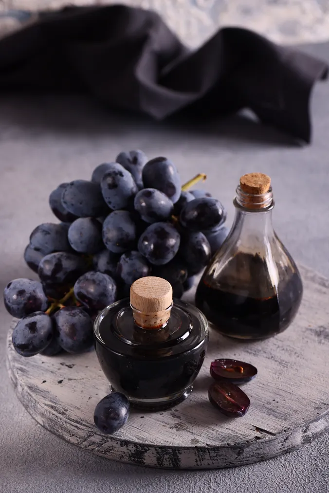 Eine Traube dunkler Trauben und zwei Flaschen Balsamico-Essig – eine runde und eine tropfenförmige – auf einer strukturierten Holzoberfläche.