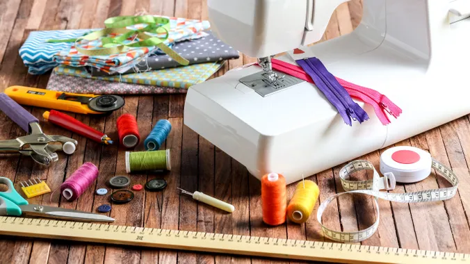 Auf einem dunklem Holztisch steht eine weiße Nähmaschine, daneben liegen einige bunte Stoffe, ein Maßband und ein Lineal, diverse Fäden und anderes Zubehör zum Nähen.