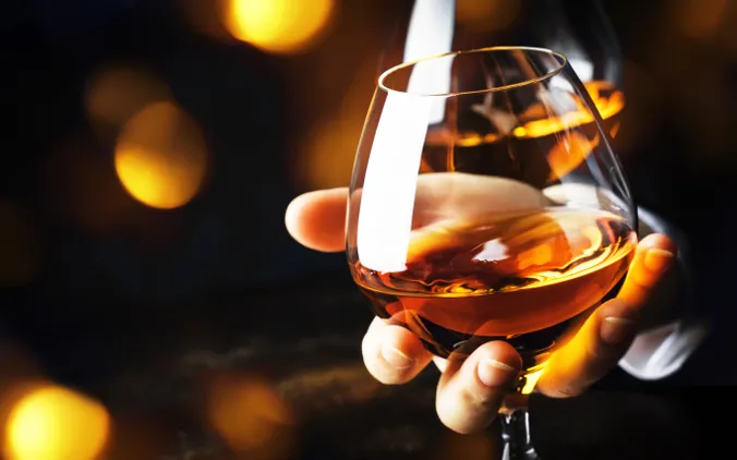 Eine Hand, die ein Cognacglas mit bernsteinfarbenem Cognac hält, im Hintergrund warme, verschwommene goldene Lichter.