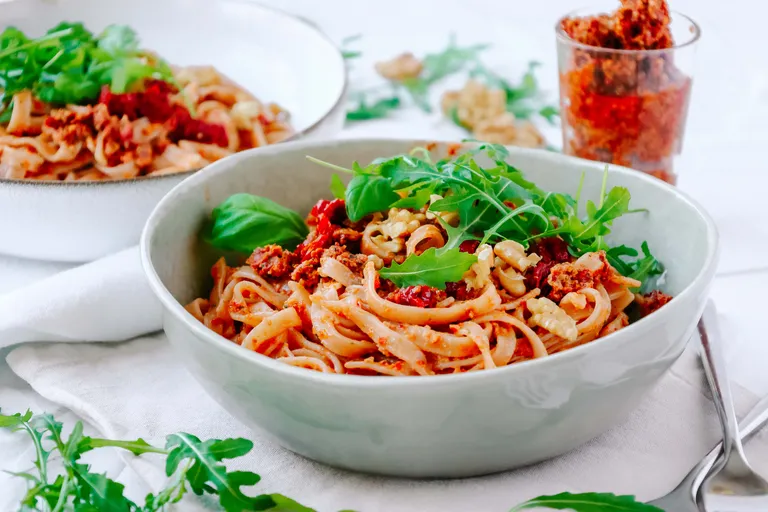 Eine Schüssel Pasta mit sonnengetrockneten Tomaten, Walnüssen und Rucola auf einem weißen Tisch, im Hintergrund eine zweite Schüssel und ein Glas mit Beilagen.