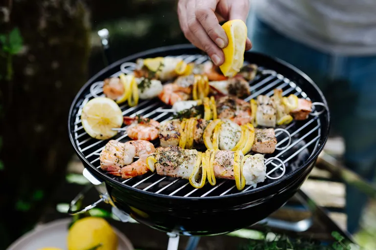 Eine Person, die auf einem Holzkohlegrill Spieße mit Meeresfrüchten und Gemüse grillt und diese mit einer Zitronenscheibe garniert.