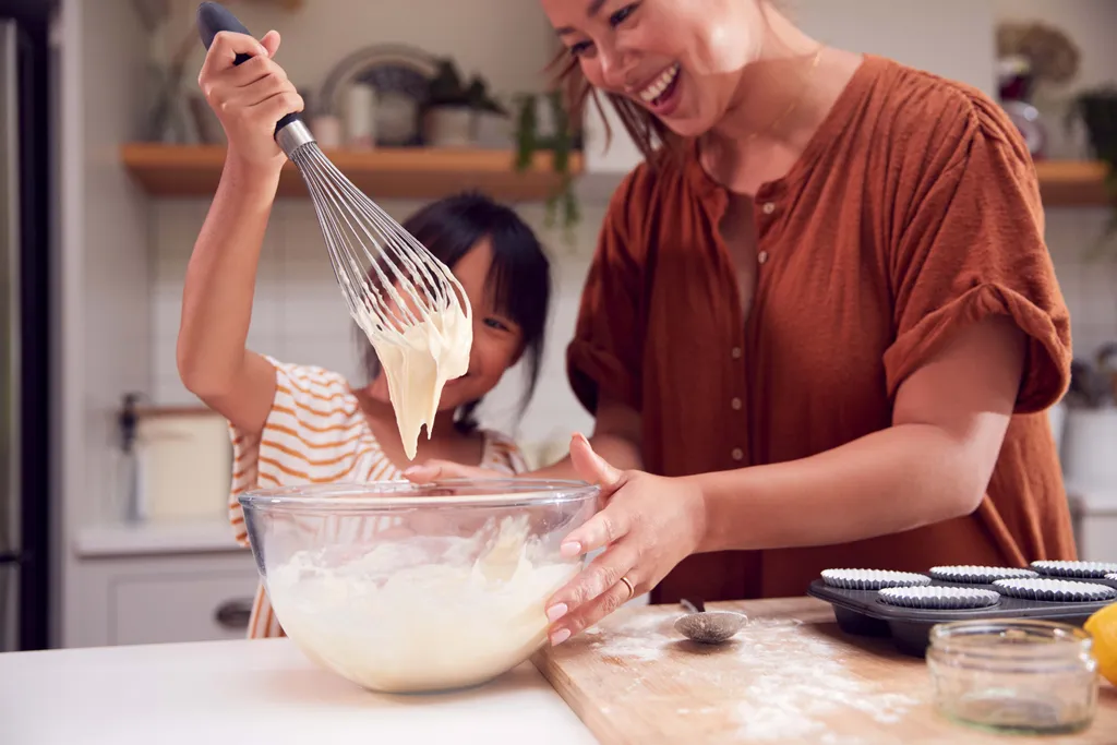 Eine lächelnde Frau und ein Kind backen gemeinsam, rühren mit einem Schneebesen Teig in einer Schüssel an und sind von Backutensilien auf einer Küchenarbeitsplatte umgeben.