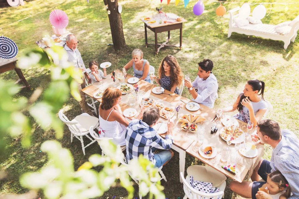 Ein Tisch bei einer Gartenparty auf dem Rasen, gedeckt mit Tischdecke und Tellern, von oben gesehen. Rundherum sitzen feiernde Personen