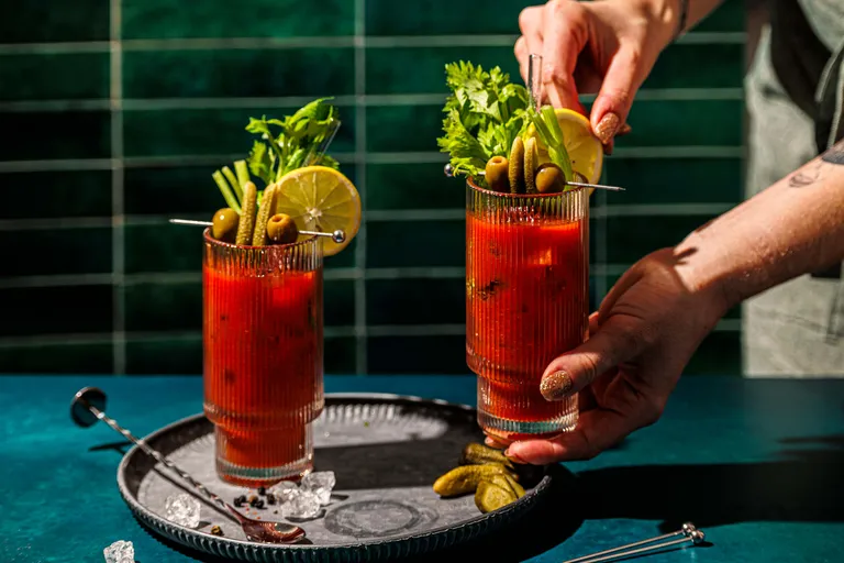 Zwei Bloody-Mary-Cocktails, garniert mit Sellerie, Oliven, Limette und Gewürzgurken, die von Händen auf einem Tablett mit Eis auf einer blaugrünen Unterlage angerichtet werden.