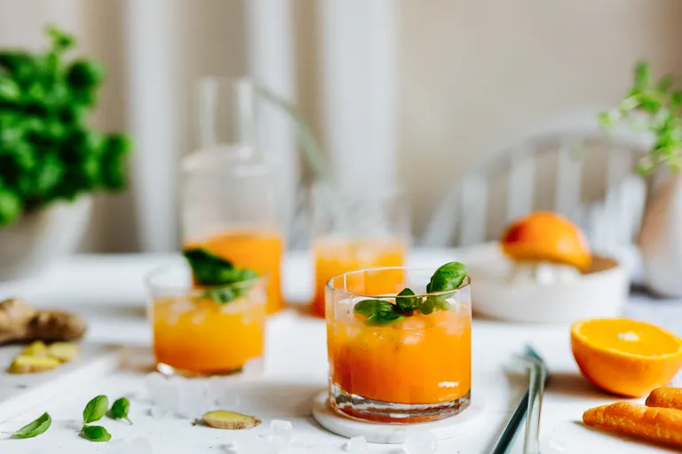 Ein Frühstückstisch mit einem Saftkrug und Gläsern gefüllt mit Marillen-Karotten-Orangen-Ingwer-Saft und dekoriert mit grünen Blättern. Im Hintergrund ist eine Orangenpresse und ein Basilikum-Stock verschwommen zu sehen.