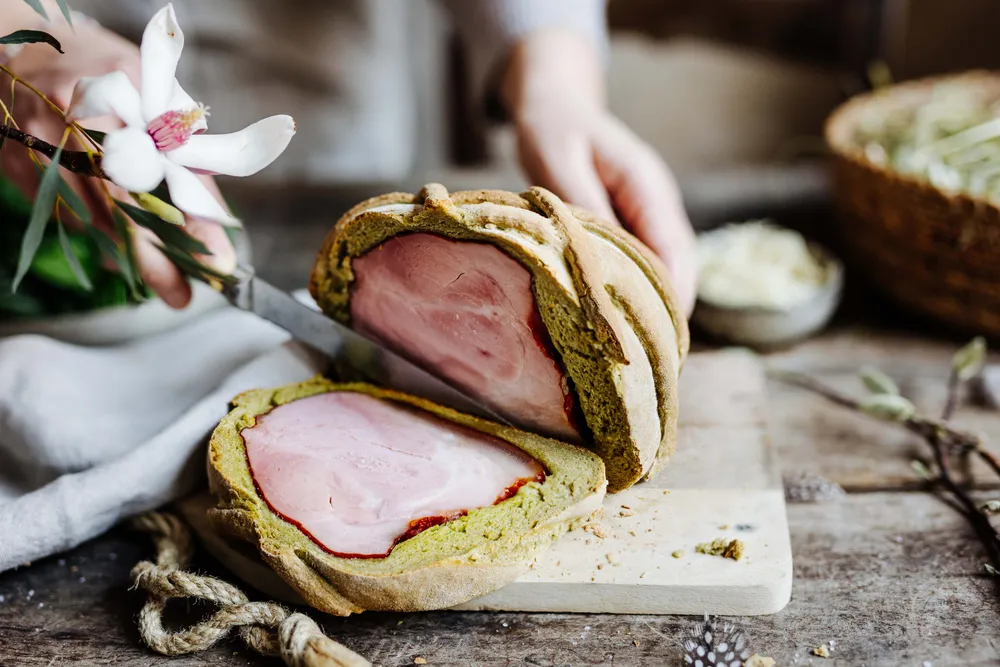 Ein Laib Brot mit Schinken in der Mitte auf einem Schneidebrett, die gerade mit einem Messer geschnitten wird; daneben liegen dekorative Blumen.