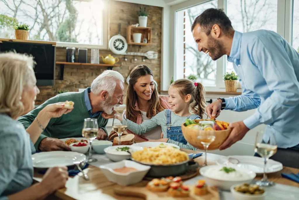 Eine Familie genießt gemeinsam ein fröhliches Essen an einem Tisch mit verschiedenen Gerichten und teilt Lachen und Geselligkeit in einem hellen Esszimmer.