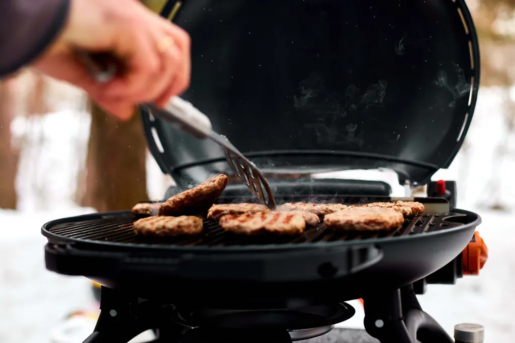 Eine Person grillt Burger auf einem Außengrill und wendet die Patties mit einer Zange. Rauch steigt vom heißen Grill auf, umgeben von einer verschneiten Landschaft.