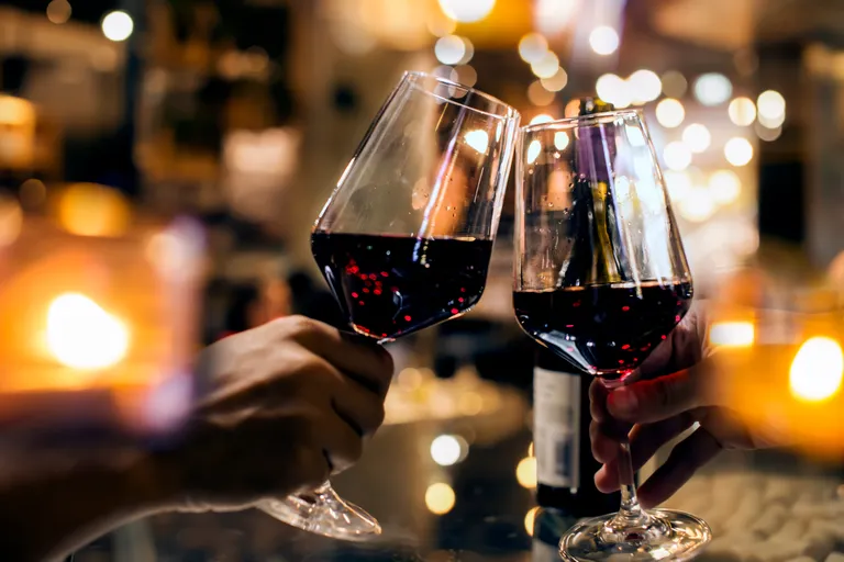 Zwei Gläser Rotwein klirren in einer warmen, sanft beleuchteten Umgebung, im Hintergrund sind verschwommene Lichter zu sehen.