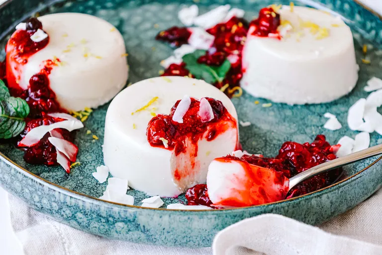 Cremige Panna Cotta mit einer Kompott aus roten Beeren und Kokosflocken, serviert auf einem blauen Teller mit einem Löffel.