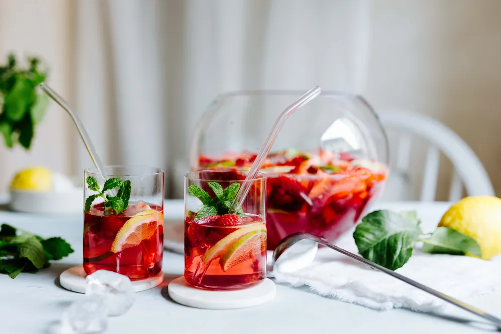 Zwei Gläser mit rotem Fruchtpunsch, garniert mit Zitronenscheiben und Minze, neben einem Krug und einer Zitrone, auf einem hellen Tisch mit verstreuten Eiswürfeln.