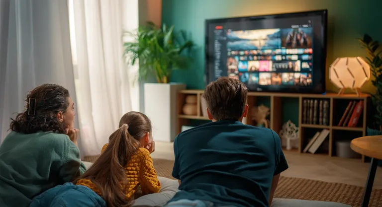Dreiköpfige Familie liegt auf der Couch im Wohnzimmer, sehen fern, neben dem Fernseher indirekte Beleuchtung