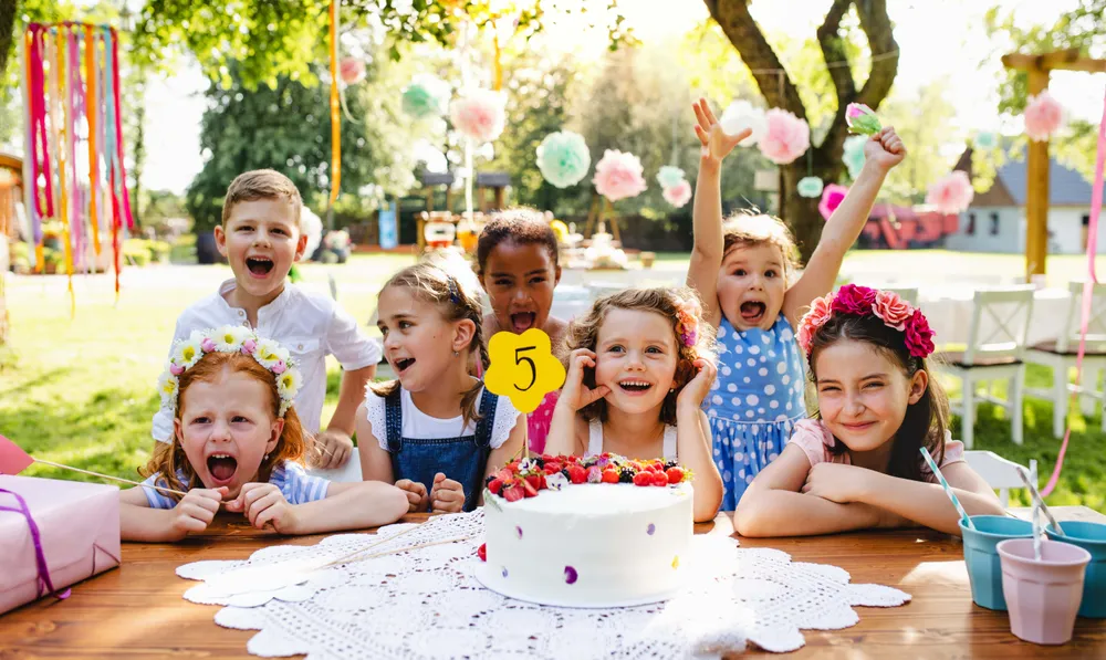 Eine Gruppe fröhlicher Kinder auf einer Geburtstagsfeier im Freien, die sich um eine Torte mit einer „5“ als Tortenfigur versammelt haben, umgeben von bunten Dekorationen.