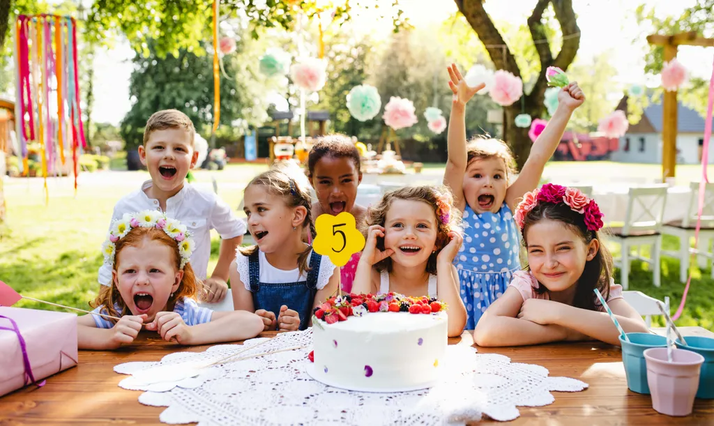 Eine Gruppe fröhlicher Kinder auf einer Geburtstagsfeier im Freien, die sich um eine Torte mit einer „5“ als Tortenfigur versammelt haben, umgeben von bunten Dekorationen.