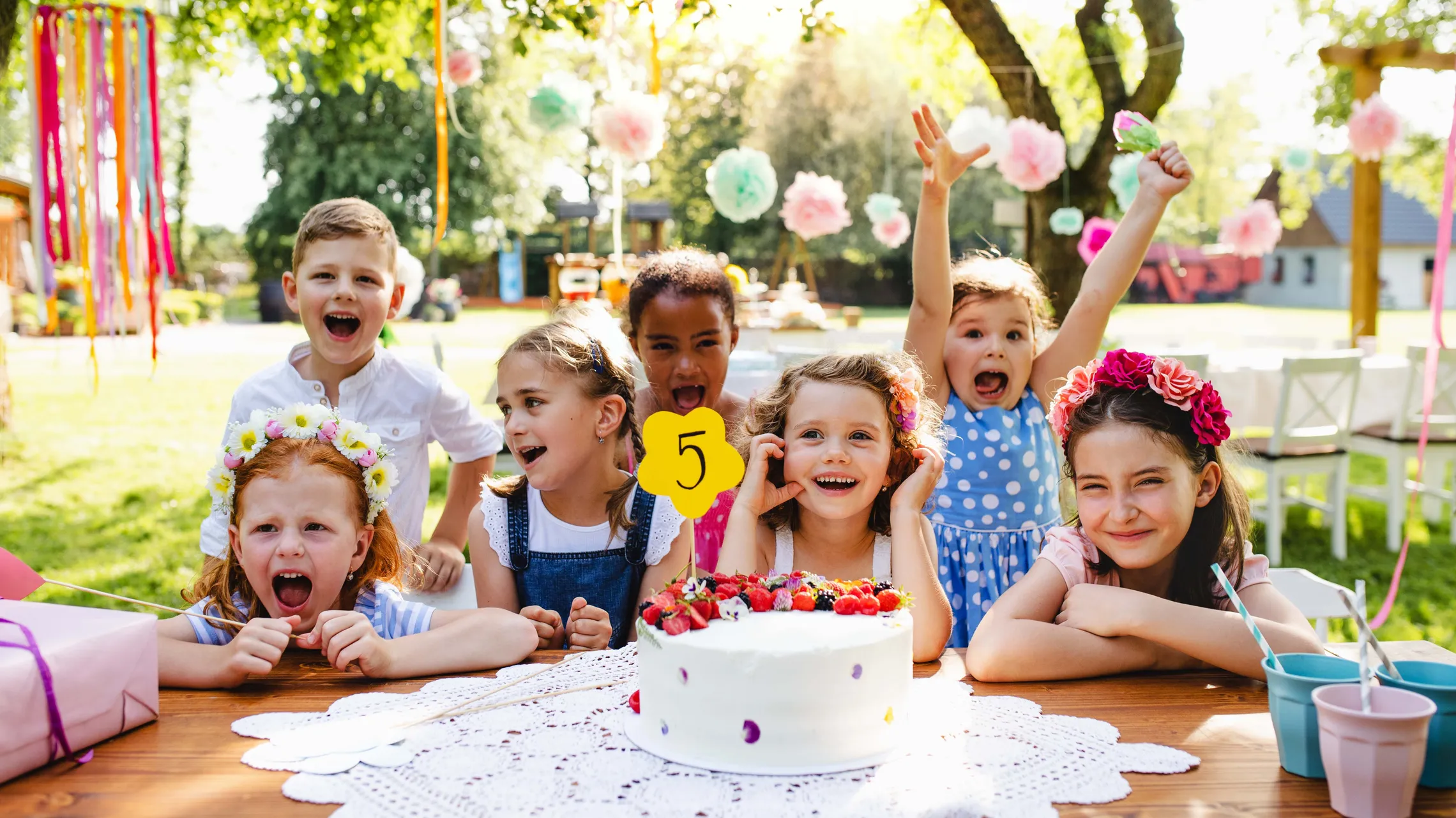 Eine Gruppe fröhlicher Kinder auf einer Geburtstagsfeier im Freien, die sich um eine Torte mit einer „5“ als Tortenfigur versammelt haben, umgeben von bunten Dekorationen.