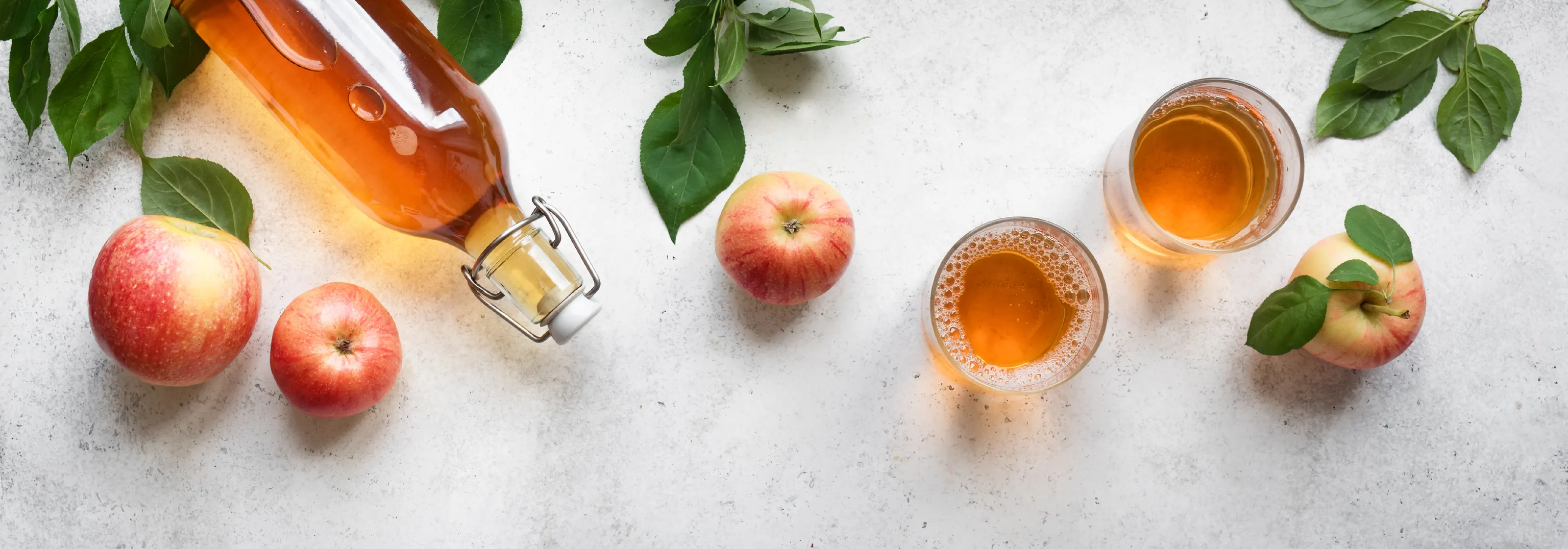 Eine Flasche und zwei Gläser Apfelwein, umgeben von frischen Äpfeln und Blättern auf einer hellen, strukturierten Oberfläche.