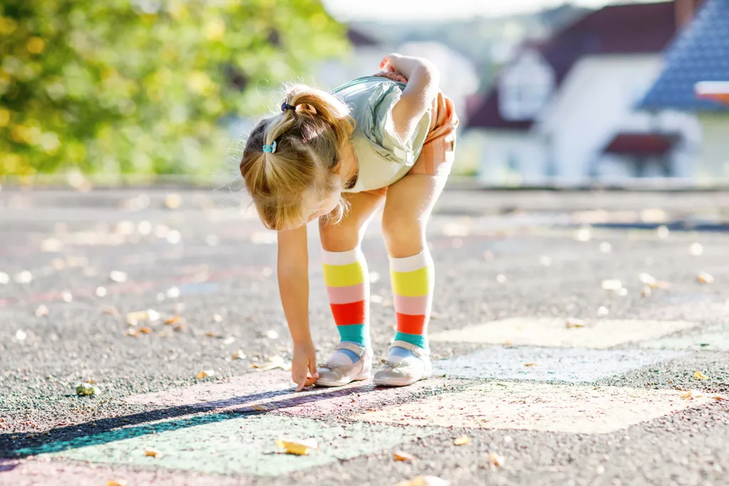 Ein Mädchen mit bunten Strümpfen malt mit Straßenkreide ein Tempelhüpf-Spiel auf Asphalt