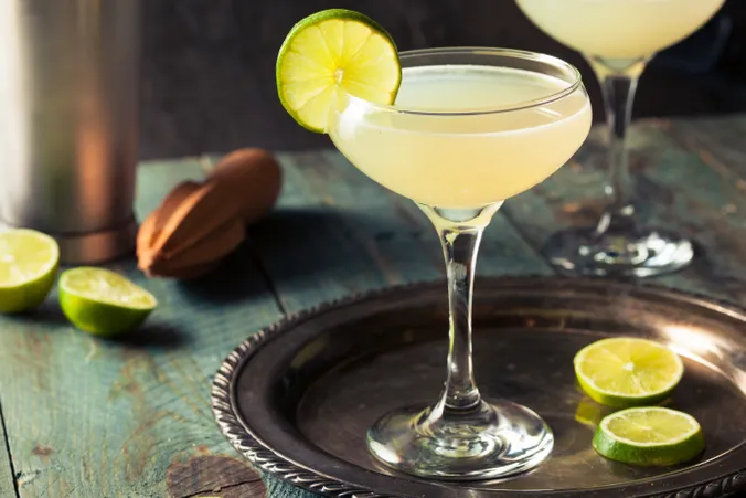 Ein Cocktail in einem Glas, garniert mit einer Limettenscheibe, steht auf einem Silbertablett, umgeben von Limettenhälften auf einem rustikalen Holztisch.