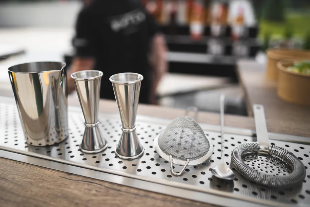 Bar-Utensilien auf einer Arbeitsplatte: Shaker, Jigger, Sieb, Löffel und Muddler auf einer perforierten Metallplatte.