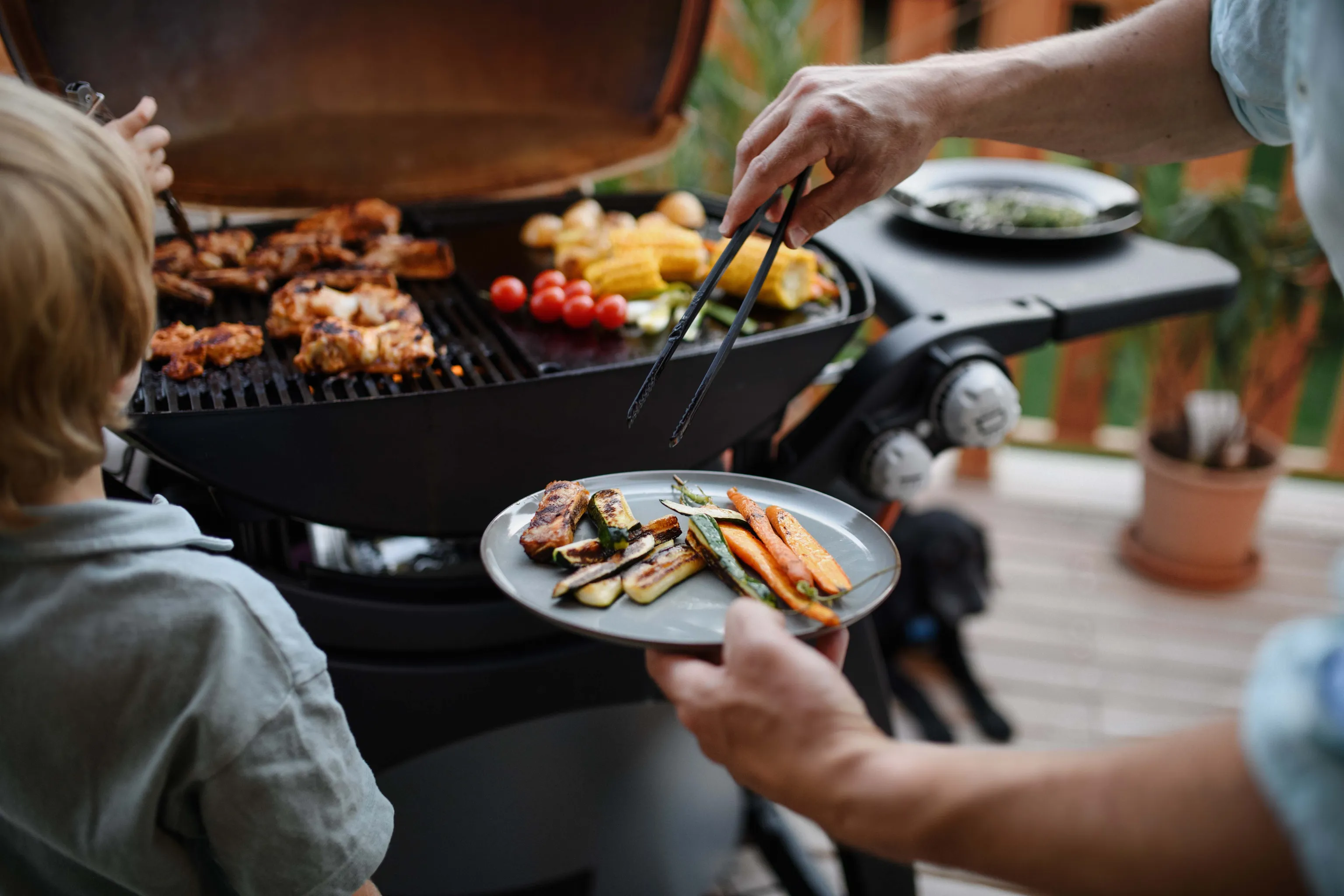 Eine Person, die auf einem Grill im Freien Fleisch und Gemüse grillt und einen Teller mit gegrillten Zucchini und Karotten in der Hand hält.