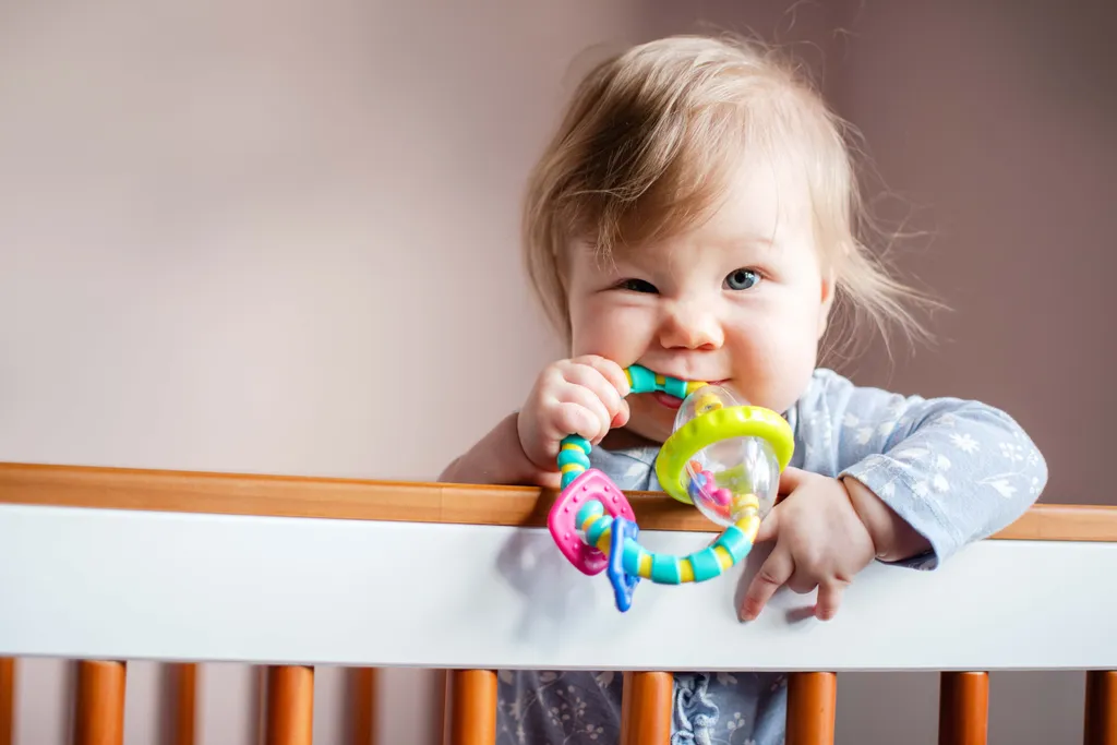 Fröhliches Baby hält einen bunten Greifling im Gitterbett und erkundet das Spielzeug