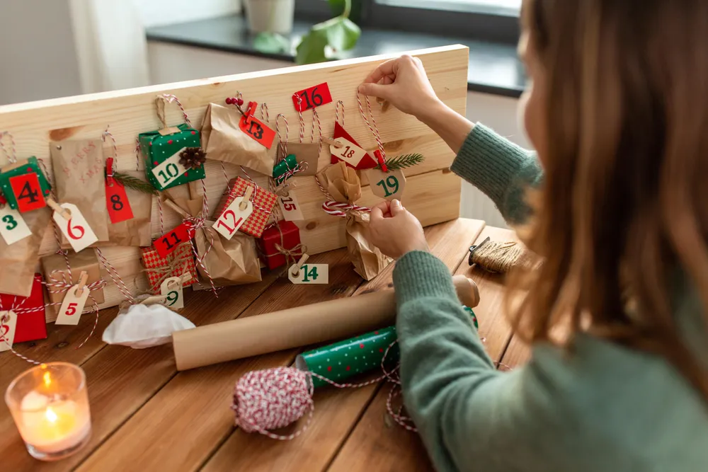 Eine Frau befüllt einen Adventkalender: Säckchen sind auf einem Holzbrett montiert, dazu rote und grüne Dekoration. Am Tisch brennt eine Kerze.