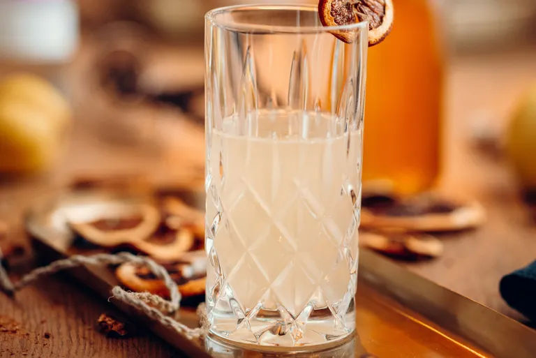 Ein Glas mit einem hellen Getränk in einem strukturierten Becher, garniert mit einer getrockneten Orangenscheibe, auf einem Tablett mit getrockneten Zitrusscheiben.