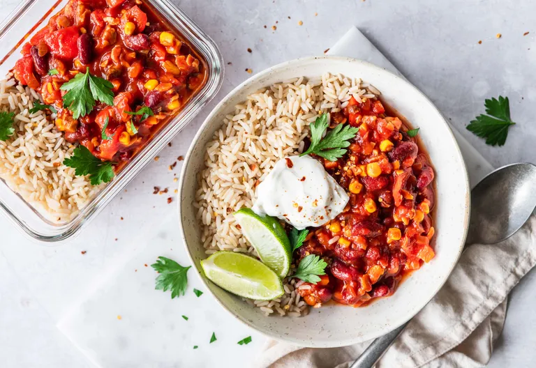 Portion Chili sin Carne mit Reis, Limette und Sauerrahm sowie Meal-Prep-Box im Hintergrund