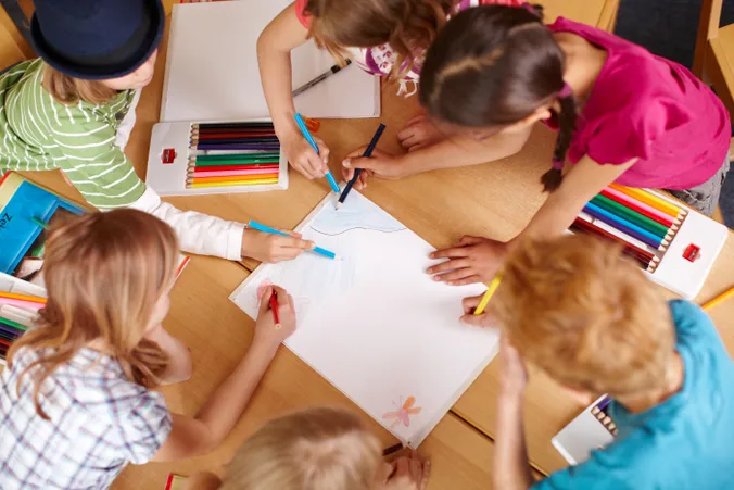 6 Kinder malen gemeinsam eine Zeichnung auf einem Blatt, auf einem Holztisch. Man sieht Buntstifte, einen Block sowie die Kinder aus der Vogelperspektive