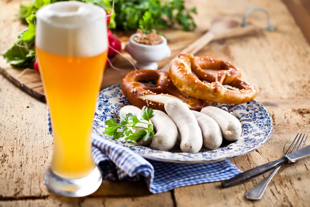 Ein großes Glas Bier, ein Teller mit Brezeln und Weißwürsten auf einem Holztisch, garniert mit Petersilie, daneben eine Serviette und eine Gabel.