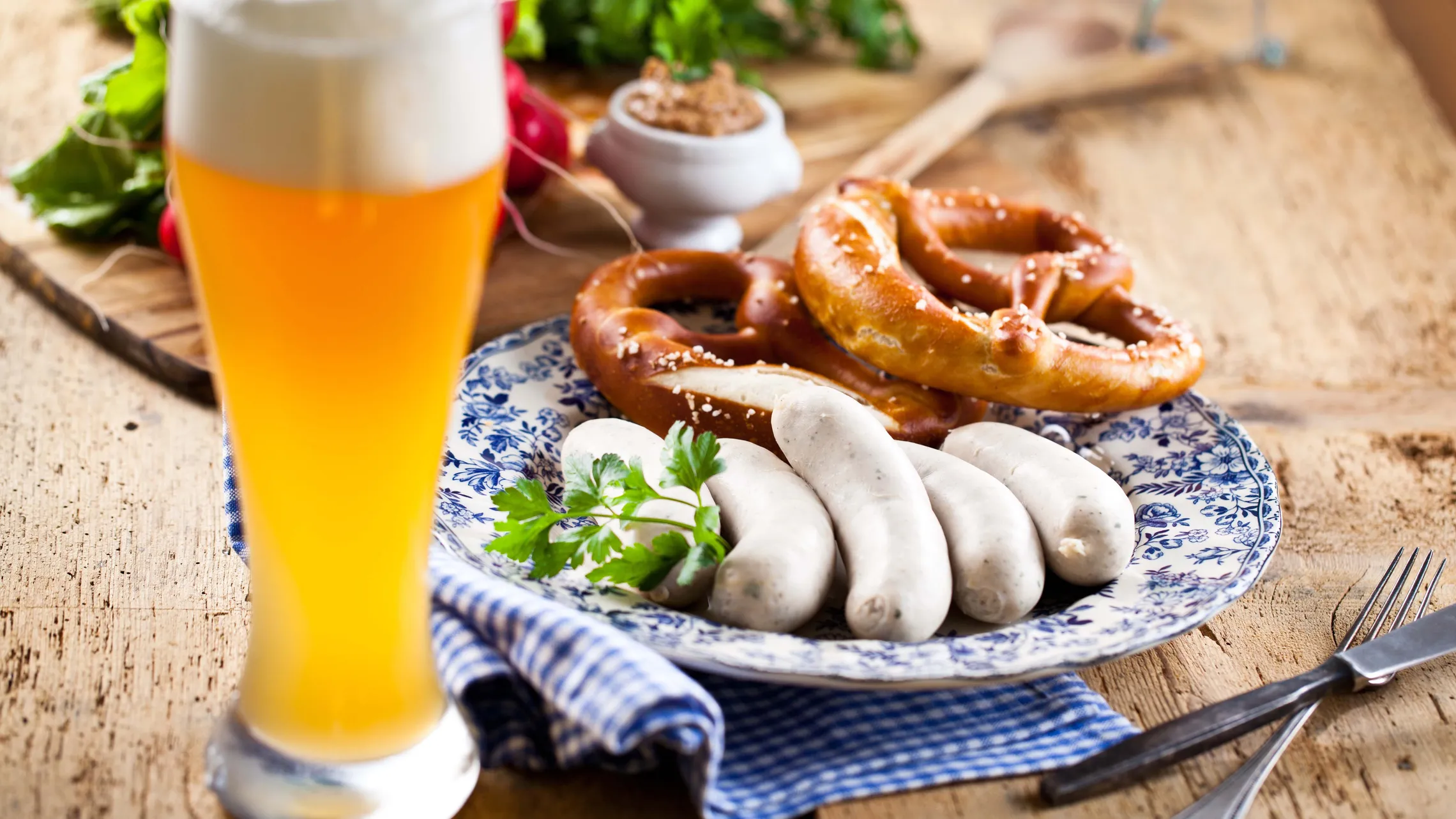 Ein großes Glas Bier, ein Teller mit Brezeln und Weißwürsten auf einem Holztisch, garniert mit Petersilie, daneben eine Serviette und eine Gabel.
