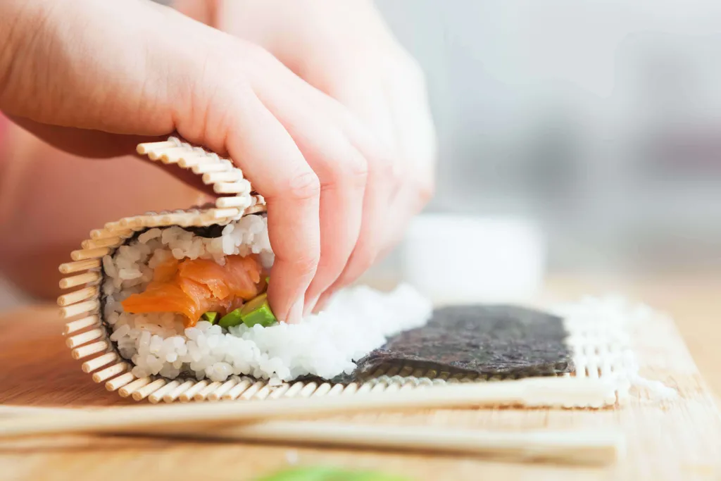 Sushi-Rolle mit Lachs und Avocado wird auf einer Bambusmatte von Hand gerollt
