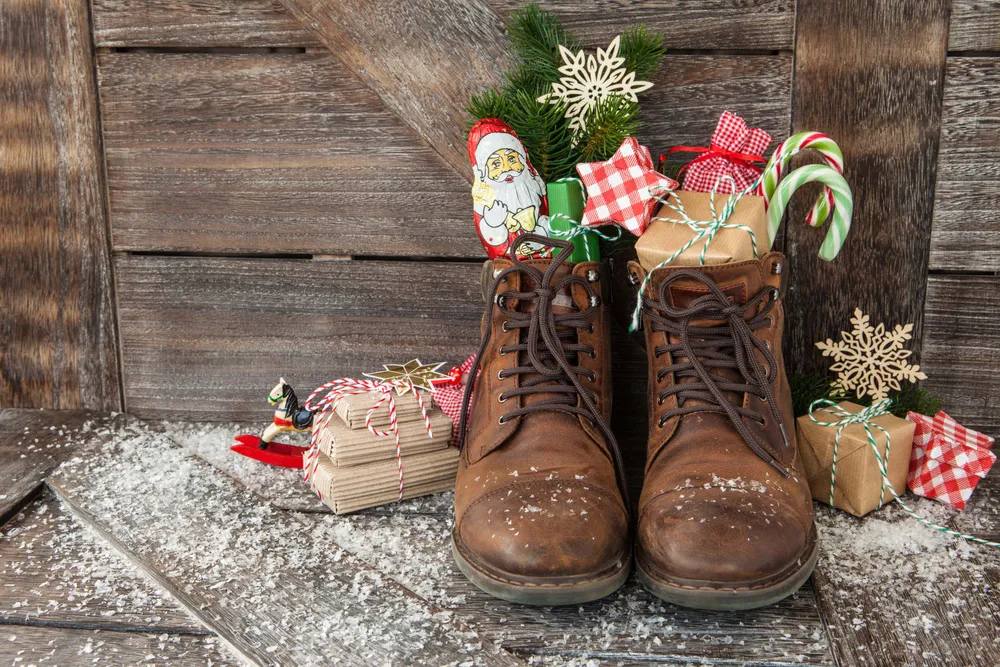 Braune Stiefel, gefüllt mit Geschenken, Zuckerstangen und Weihnachtsschmuck, umgeben von verpackten Geschenken und Kunstschnee auf einem Holzboden.