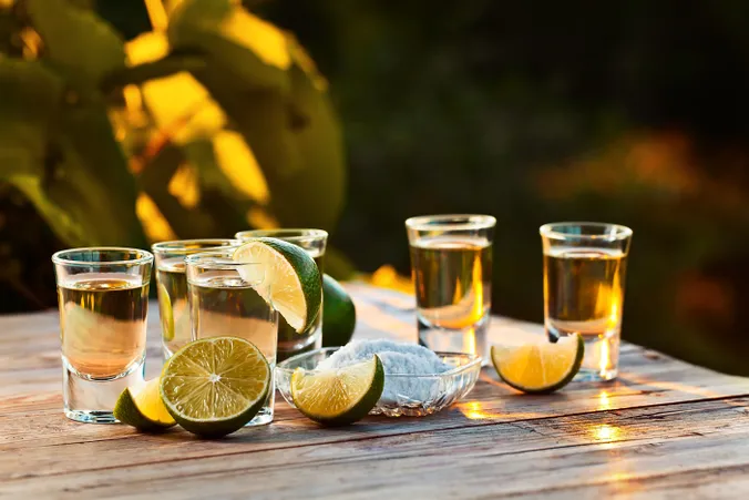 Fünf Tequila-Shots mit Limettenschnitzen und einer Schale mit Salz auf einem Holztisch, der im warmen Licht des Sonnenuntergangs erstrahlt.